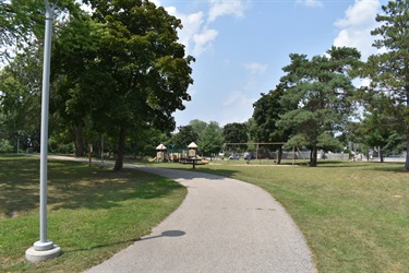A paved walking path curves through a grassy park with trees and a playground in the background.