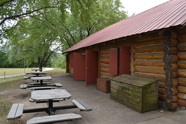 A wooden park shelter building with a red roof and picnic tables outside.