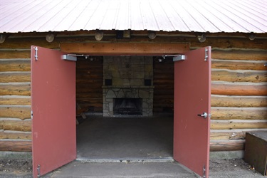 A rustic park shelter building with a large stone fireplace visible through open red double doors.
