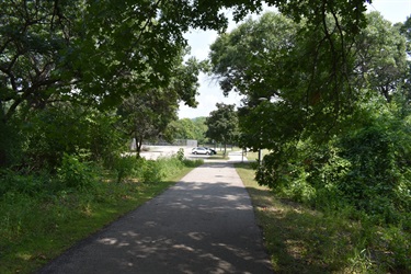 A paved walking path leads through a park with lush green trees on either side. In the distance, a white car is parked near a building.