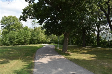 A paved walking path curves through a park with trees and grass on either side under a partly cloudy sky.