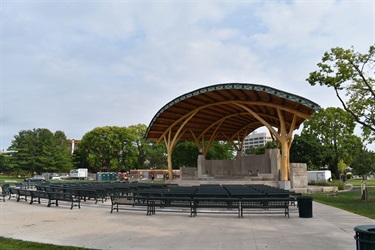 An outdoor amphitheater with a wooden canopy structure and rows of empty green benches in front of the stage.