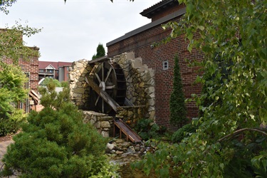 A rustic stone structure with a wooden water wheel next to a brick building, surrounded by greenery.