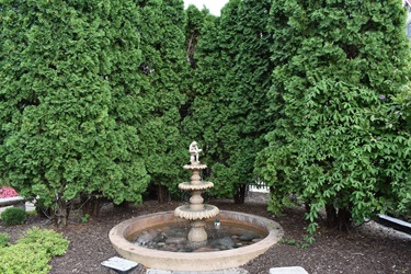 A tiered stone fountain with a statue on top sits in a circular basin filled with water and surrounded by lush green trees and bushes.