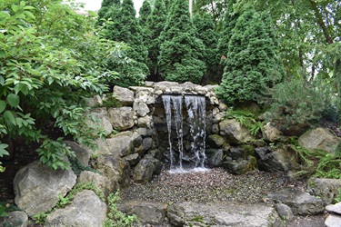 A small, multi-tiered waterfall cascades over rocks into a gravel basin, surrounded by lush green foliage and tall evergreen trees.