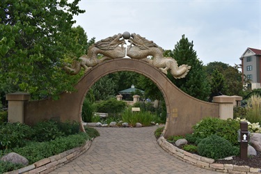 A stone archway featuring two dragon sculptures facing each other over a sphere, leading to a garden with a gazebo and lush greenery.
