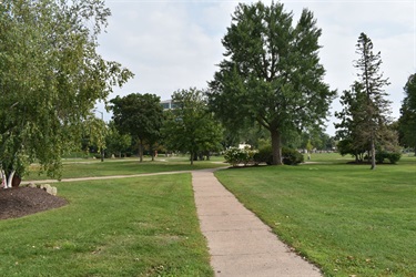 A paved pathway curves through a grassy park with various trees and a cloudy sky overhead.
