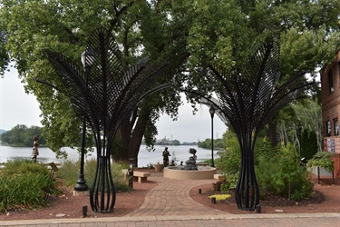 Two large black metal palm trees frame a view of a fountain and sculptures in a park by a river.