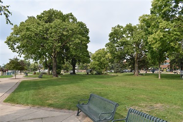 A paved walkway leads into a grassy park with mature trees and benches. In the foreground, two green metal benches are visible.