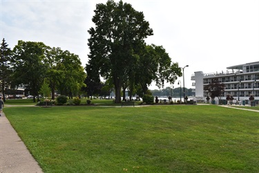 A large green lawn with a paved walkway on the left, leading to a body of water with a large cruise ship docked on the right shore, and trees scattered throughout the park.