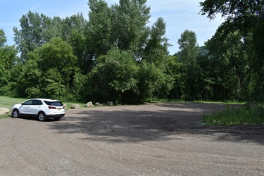 A white SUV is parked in a gravel parking lot surrounded by lush green trees and foliage under a bright blue sky.