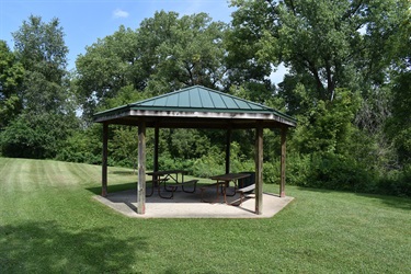 A hexagonal pavilion with a green roof and wooden supports stands in a grassy park area with picnic tables inside.
