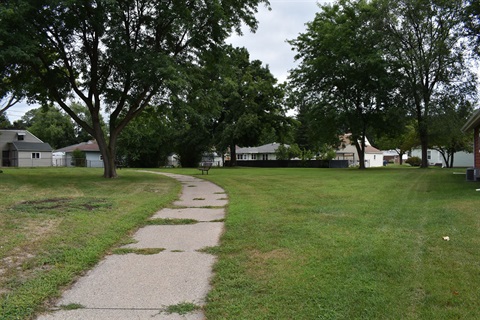 A cracked concrete path curves through a grassy park with mature trees and houses in the background under a cloudy sky.