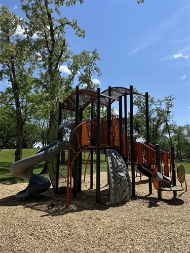 A colorful playground structure with slides, climbing walls, and bridges, set on a wood chip ground with trees and a blue sky in the background.