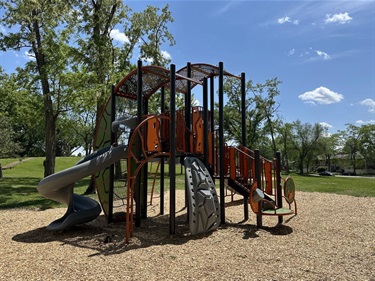 A modern playground structure with orange and gray slides and climbing elements, set on a wood chip surface under a bright blue sky with scattered clouds.