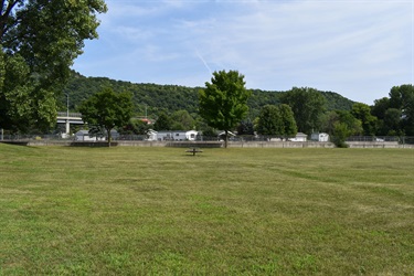 A wide grassy field with trees in the background and a hill covered in trees under a blue sky with scattered clouds.