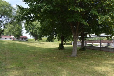 A grassy lawn slopes down towards a river with a bridge in the background, surrounded by trees and a house on the left.
