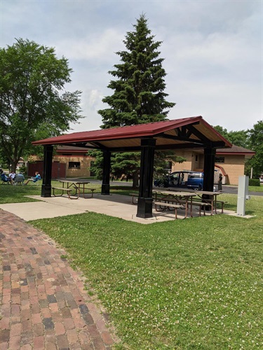 A covered picnic pavilion with picnic tables and benches is set in a grassy park area with a brick walkway.