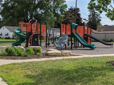 A modern playground with orange and green structures, including slides, climbing walls, and swings, set against a backdrop of trees and houses.