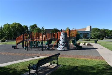 A modern playground with colorful slides, climbing structures, and swings, set against a backdrop of trees and a building under a clear blue sky. A bench sits in the foreground.