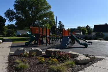 A modern playground with green and orange slides, a climbing wall, and a rope net structure on a paved surface with surrounding trees and houses.