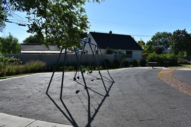 A swing set stands on a playground with a house and trees in the background on a sunny day.