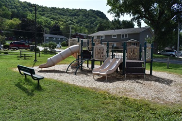 A playground with two slides, a climbing structure, and a bench sits on wood chips surrounded by grass. In the background are apartment buildings and a tree-covered hill.