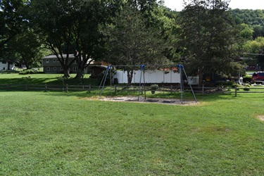 A swing set with three seats stands in a grassy area with trees and houses in the background.