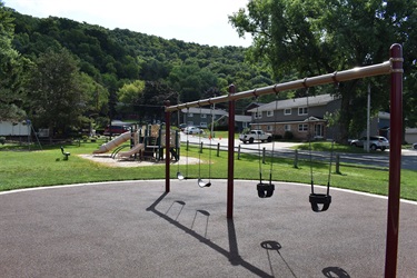 A playground with swings in the foreground and a jungle gym with slides in the background, set against a backdrop of trees and houses.
