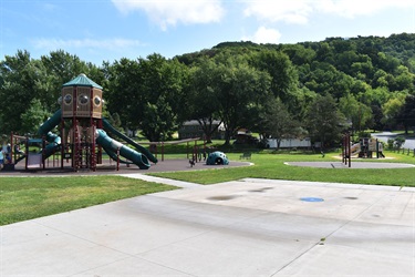 A playground with a large climbing structure and slides, surrounded by green grass and trees with a hill in the background.