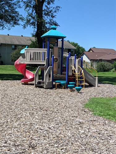 A colorful playground structure with slides, a climbing wall, and platforms, set on a wood chip surface with grass and buildings in the background.