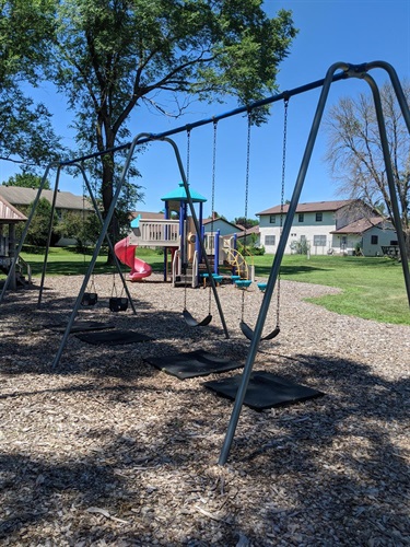 A playground with swings in the foreground and a colorful play structure with a red slide in the background, set against a backdrop of trees and houses on a sunny day.