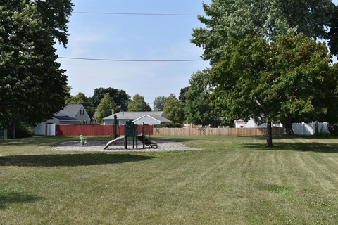 A small playground with a slide and a seesaw is visible in a grassy park area with large trees and houses in the background.