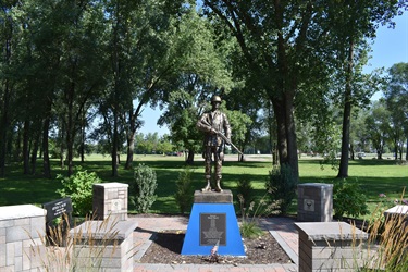 A paved walkway leads to a statue of a soldier on a blue base, flanked by stone pillars and lush greenery.