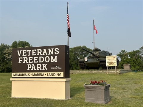 A sign for Veterans Freedom Park with a tank in the background under a clear sky.