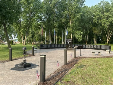 A Vietnam War memorial with a curved wall inscribed with names, a helmet and boots monument, and flags flying in a park setting.