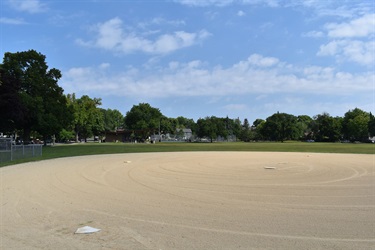 A baseball field with dirt infield and green outfield under a blue sky with scattered clouds. Trees line the background behind the field.