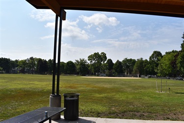 View of a baseball field from under a pavilion with a picnic table and trash can in the foreground.