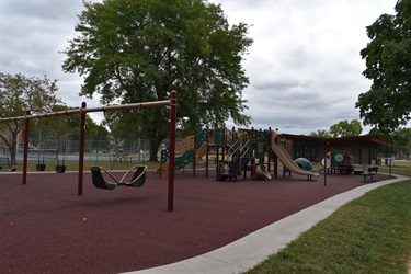 A modern playground with swings, slides, and climbing structures, set against a backdrop of trees and a cloudy sky.