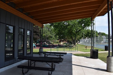 A covered patio with picnic tables overlooks a park with a playground and tennis courts.