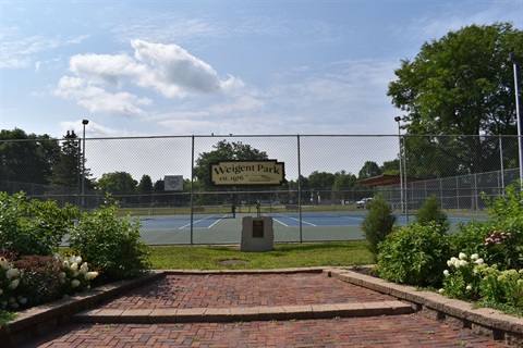 A view of a tennis court at Weigent Park, established in 1976, with a brick pathway in the foreground and lush greenery on either side.