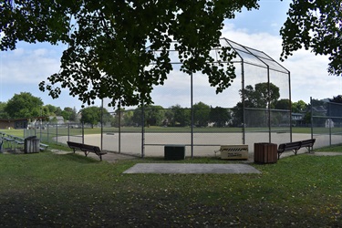 A baseball field with a backstop and bleachers under a cloudy sky, viewed from a grassy area with trees.
