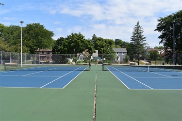 Two empty blue tennis courts with green out-of-bounds areas are shown on a sunny day with scattered clouds.