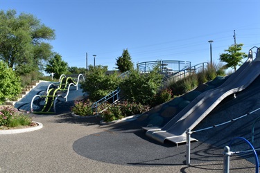 A modern playground with metal slides, climbing structures, and landscaping under a clear blue sky.