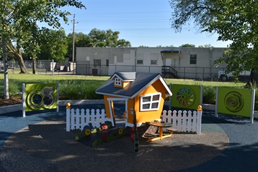 A vibrant orange playhouse with white trim and a picnic table inside is surrounded by a white picket fence. Green panels with colorful circular designs are on either side of the playhouse.