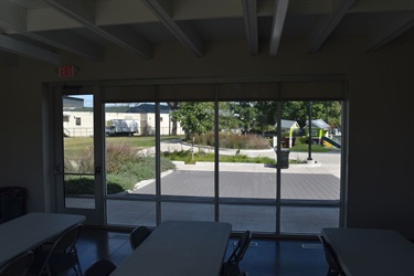 View from inside a room with tables, looking out a large window to a park with a playground and buildings beyond.