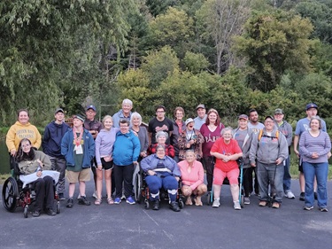 A group of people, some in wheelchairs, pose for a photo outdoors with trees and foliage in the background.