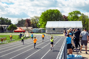 Kids run and play on an outdoor school track as spectators line the fence; under a blue sky.