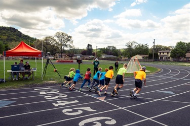 A youth track race on an outdoor track with runners in colorful shirts sprinting past an orange cone and a red tent at the side.