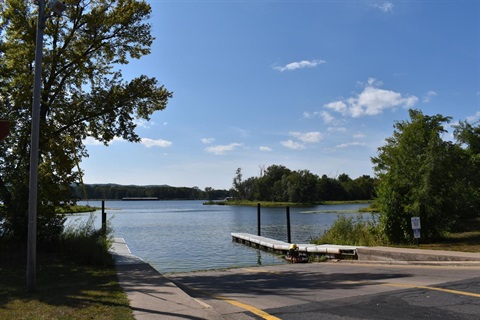 A concrete boat ramp leads into a wide river under a blue sky with scattered clouds. A floating dock is on the right side of the ramp.